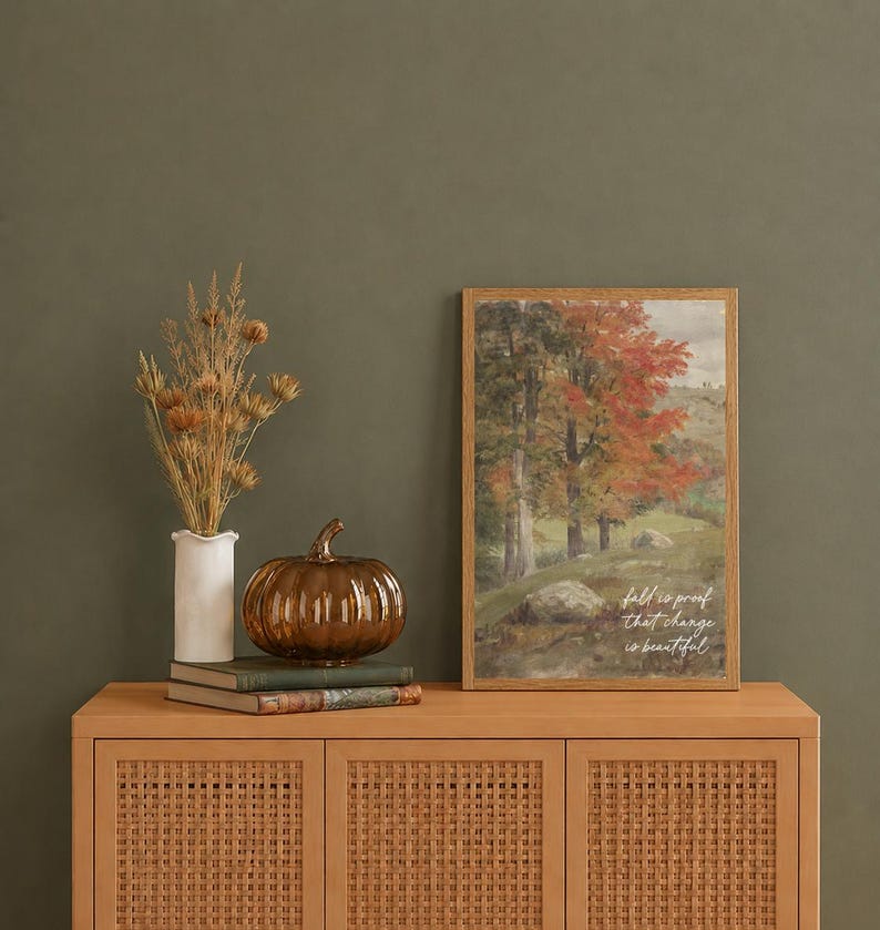 May include: A framed vertical print featuring a landscape painting of autumn trees with red and green foliage, including the text "fall is proof that change is beautiful". Beside it sits a white vase with dried grasses and a brown glass pumpkin.