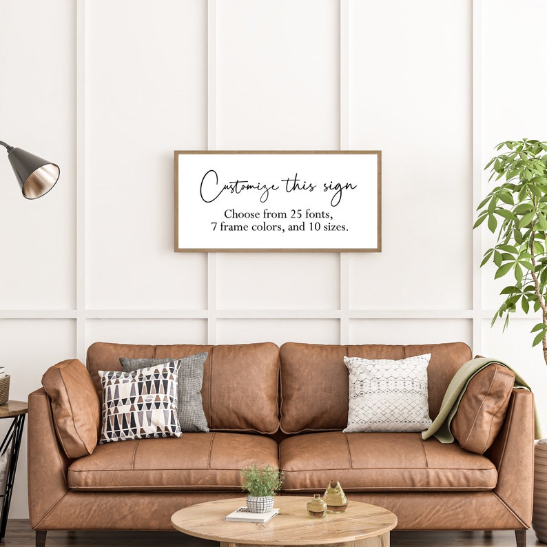 Pu&ograve; includere: Un cartello da parete rettangolare con una sottile cornice in legno che presenta il testo nero "Customize this sign" seguito da "Choose from 25 fonts, 7 frame colors, and 10 sizes." Il cartello &egrave; esposto su una parete bianca sopra un divano in pelle marrone.