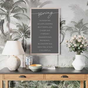 May include: A framed print with the word "spring" in cursive script at the top, followed by a list of spring-related words in white. The print is displayed on a dark grey background. The frame is light-coloured wood. The background is a tropical palm tree wallpaper.