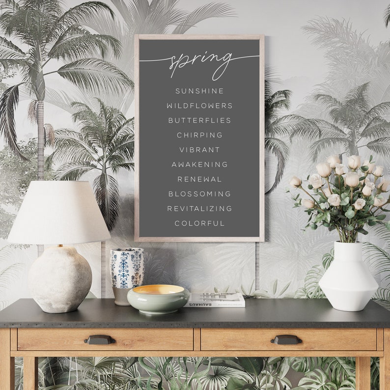 May include: A framed print with the word "spring" in cursive script at the top, followed by a list of spring-related words in white. The print is displayed on a dark grey background. The frame is light-coloured wood. The background is a tropical palm tree wallpaper.