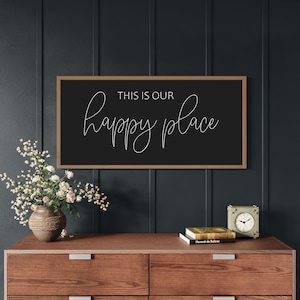 May include: A rectangular wall sign with a thin wooden frame and a black background. The text "THIS IS OUR" is printed in a simple sans-serif font above the word "happy place" written in a white, flowing, cursive script lettering style.