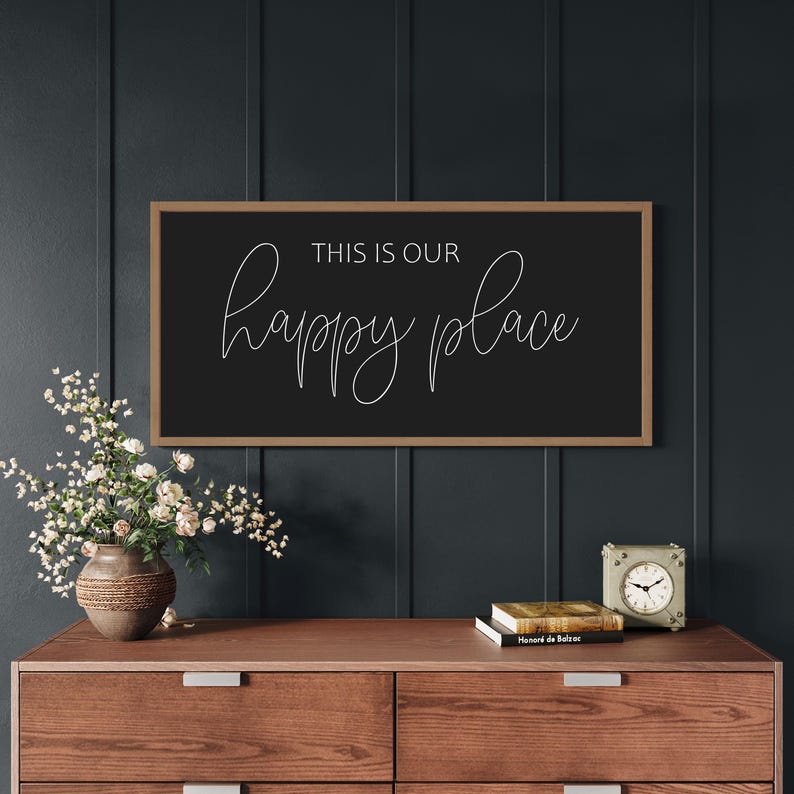 May include: A rectangular wall sign with a thin wooden frame and a black background. The text "THIS IS OUR" is printed in a simple sans-serif font above the word "happy place" written in a white, flowing, cursive script lettering style.