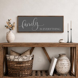 May include: A rectangular wall sign with a dark gray background and a wooden frame. The text "family IS EVERYTHING" is printed in white, with the word "family" in a cursive script font and "IS EVERYTHING" in a clean, uppercase sans-serif font.