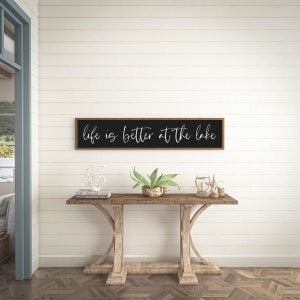 May include: A rectangular wall sign with a dark frame and black background featuring the white cursive text "life is better at the lake" centered on the surface. The sign is mounted horizontally on a white horizontal plank wall.