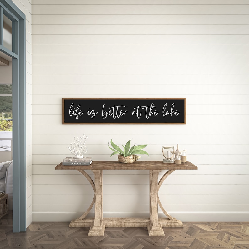 May include: A rectangular wall sign with a dark frame and black background featuring the white cursive text "life is better at the lake" centered on the surface. The sign is mounted horizontally on a white horizontal plank wall.