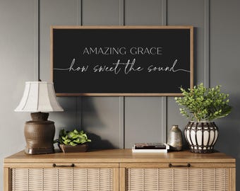 Puede incluir: Un letrero de pared rectangular con fondo negro y marco de madera clara. El texto "AMAZING GRACE" está impreso en una fuente sans-serif sobre el texto en cursiva "how sweet the sound" en blanco, centrado en el letrero.