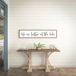 May include: A rectangular wall sign with a thin brown wooden frame and a white background featuring the black cursive text "life is better at the lake" centered on the surface. The sign is mounted horizontally on a white horizontal plank wall.