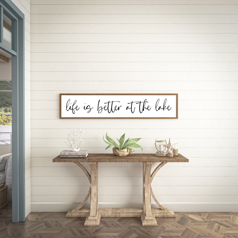 May include: A rectangular wall sign with a thin brown wooden frame and a white background featuring the black cursive text "life is better at the lake" centered on the surface. The sign is mounted horizontally on a white horizontal plank wall.