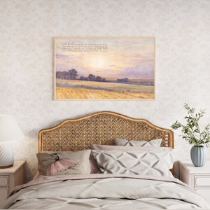 A framed print with a watercolor painting of a field with a sunset in the background. The print has the text "Seek First" and a Bible verse from Matthew 6:33. The print is hanging above a wicker headboard on a bed with a white comforter and pillows.
