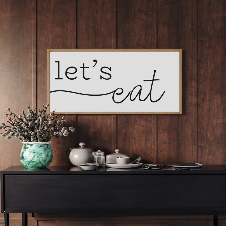 May include: A rectangular wooden sign with the words "let's eat" in black script. The sign is mounted on a dark wood-paneled wall above a black cabinet. A vase with greenery, a teapot, and dishes are on the cabinet.
