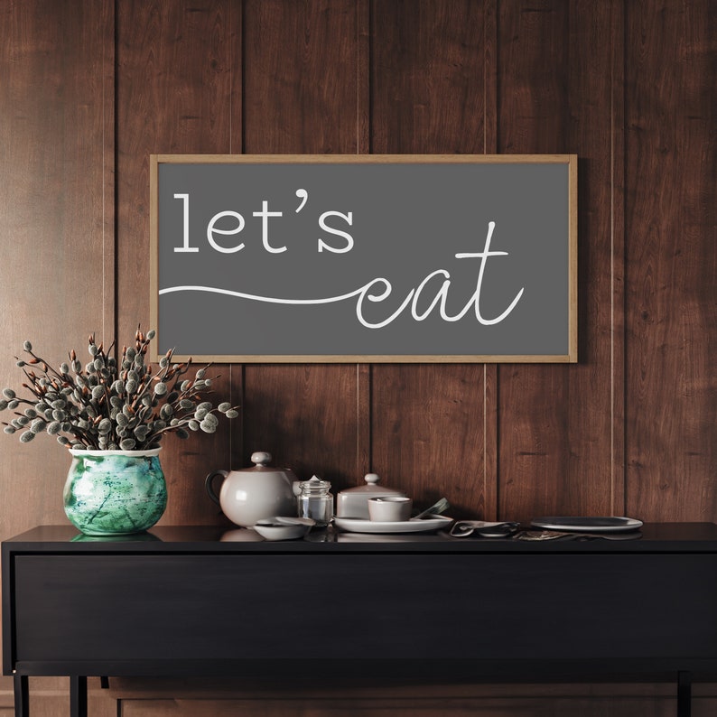 May include: A framed sign with the words "let's eat" in white script on a gray background. The sign is mounted on a dark wood-paneled wall above a black cabinet. A vase with green and blue accents and a teapot are on the cabinet.