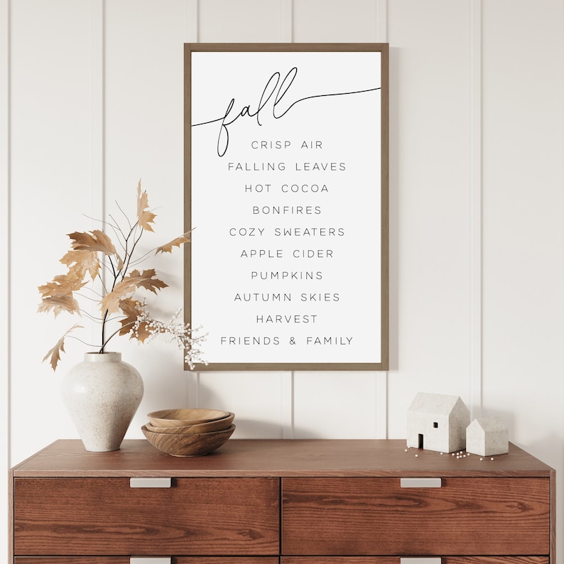 Op de afbeelding: Een ingelijst print met het woord "herfst" in cursief schrift en een lijst met herfstgerelateerde woorden, waaronder "frisse lucht", "vallende bladeren", "warme chocolademelk", "vuurkorven", "gezellige truien", "appelcider", "pompoenen", "herfstlucht", "oogst" en "vrienden en familie".
