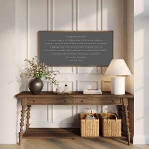 May include: A wooden console table with a framed quote, a lamp, and decorative items. The dark gray framed quote reads "So laugh often and much...This is to have succeeded." Two woven baskets sit below the table.