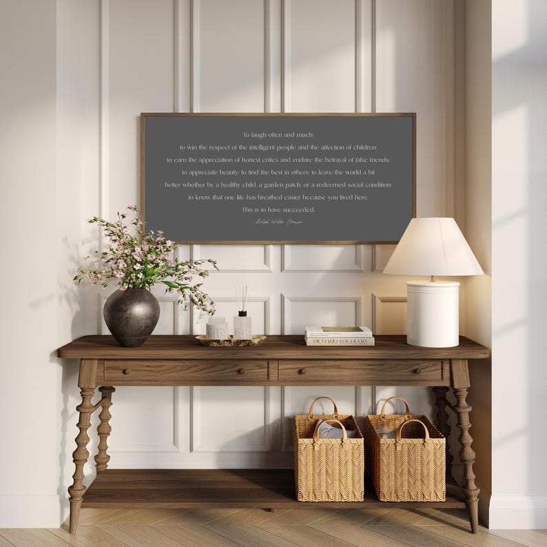 May include: A wooden console table with a framed quote, a lamp, and decorative items. The dark gray framed quote reads "So laugh often and much...This is to have succeeded." Two woven baskets sit below the table.