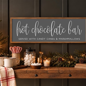 May include: A rectangular wooden-framed sign with a dark grey background and white text that reads "hot chocolate bar" in script and "SERVED WITH CANDY CANES & MARSHMALLOWS" in block letters, mounted on a dark panelled wall.