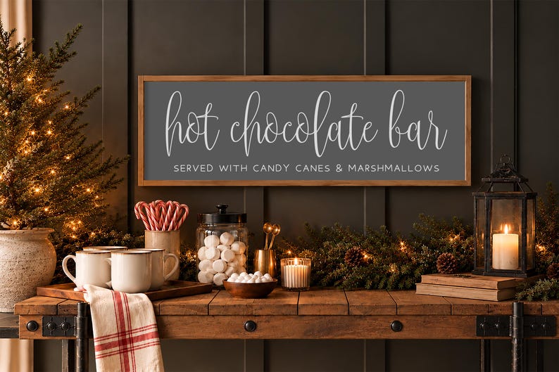 May include: A rectangular wooden-framed sign with a dark grey background and white text that reads "hot chocolate bar" in script and "SERVED WITH CANDY CANES & MARSHMALLOWS" in block letters, mounted on a dark panelled wall.