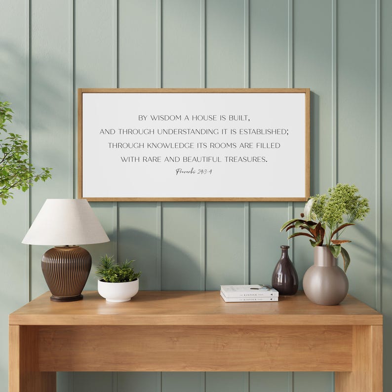 May include: A rectangular wall sign with a light wood frame and a white background featuring black text that reads: By wisdom a house is built, and through understanding it is established; through knowledge its rooms are filled with rare and beautiful treasures. Proverbs 24:3-4.