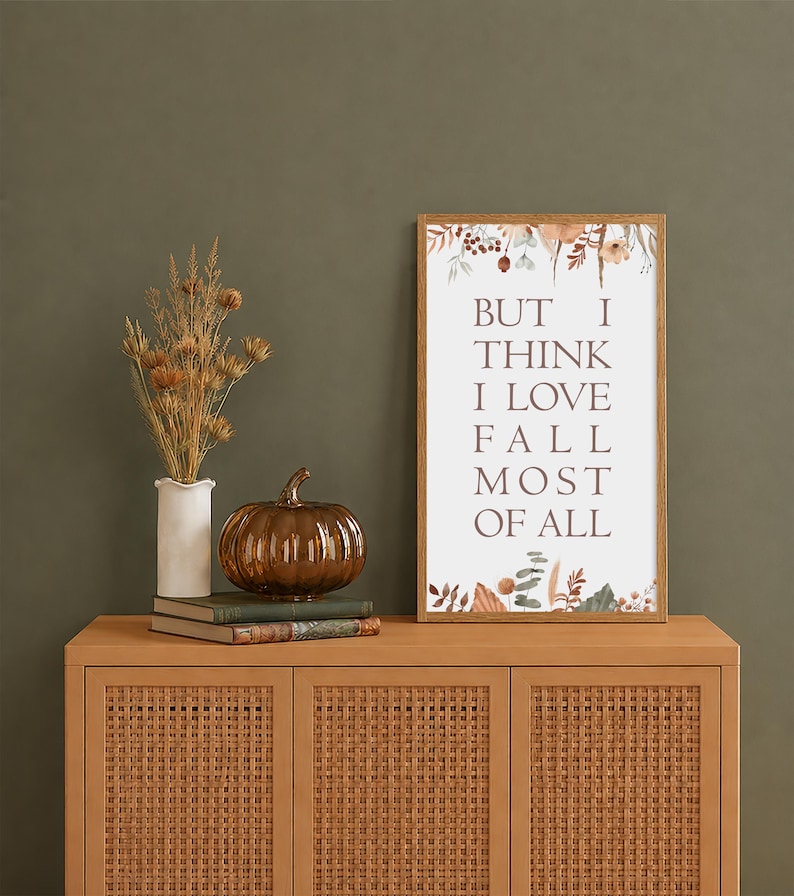 May include: A rectangular framed wall art print featuring the text BUT I THINK I LOVE FALL MOST OF ALL in dark brown lettering, decorated with botanical illustrations of dried leaves and flowers in muted autumn tones at the top and bottom.