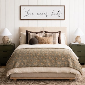 May include: A bedroom scene featuring a bed with a beige headboard, layered with pillows and a patterned blanket. Above the bed, a framed sign reads "Love never fails." Two green nightstands with lamps flank the bed, set on a patterned rug.