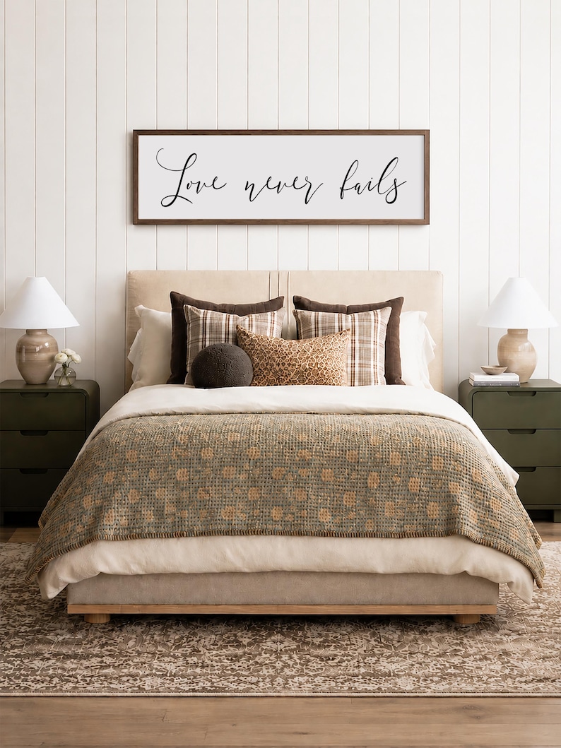 May include: A bedroom scene featuring a bed with a beige headboard, layered with pillows and a patterned blanket. Above the bed, a framed sign reads "Love never fails." Two green nightstands with lamps flank the bed, set on a patterned rug.