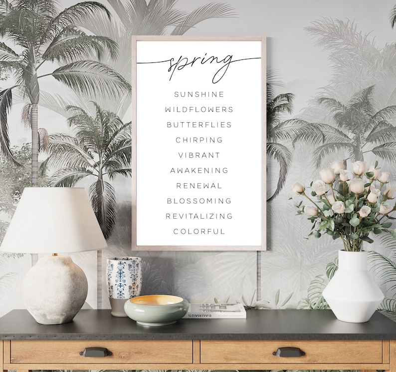 May include: A framed print with the word "spring" in cursive, followed by a list of spring-related words. The print is displayed on a dark wooden table with a lamp, vase, and decorative items. The background features a tropical palm tree wallpaper.