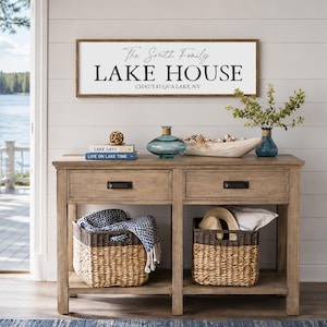 Puede incluir: Un letrero rectangular de madera con fondo blanco y texto negro que dice The Smith Family LAKE HOUSE CHAUTAUQUA LAKE, NY, montado sobre una mesa consola de madera de color marrón claro con dos cajones y dos cestas tejidas en el estante inferior.