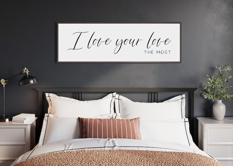 May include: A black framed sign with white background and the text "I love your love the most" in black cursive font, hanging above a bed with white bedding and a brown and white striped throw pillow.