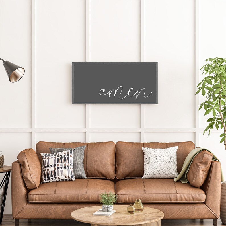 May include: A rectangular wall art piece featuring a dark gray background with the word amen written in white cursive script. The artwork is framed in a thin dark border and is mounted on a white wall above a brown leather sofa.
