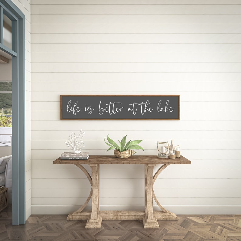 May include: A rectangular wall sign with a dark gray background and a thin brown wooden frame. The text "life is better at the lake" is written in white cursive script across the center of the sign. The sign is mounted on a white wall.