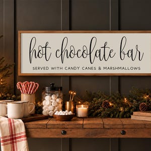 May include: A rectangular wooden sign with a light background and a dark wood frame, featuring the black text "hot chocolate bar" in a script font and "SERVED WITH CANDY CANES & MARSHMALLOWS" in a simple sans-serif font.