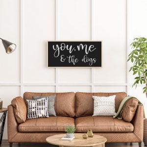 May include: A rectangular wall sign with a light wood frame and a black background featuring the white cursive text "you, me & the dogs" centered on the surface. The sign is mounted horizontally on a white wall above a brown leather sofa.