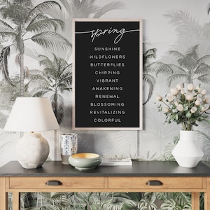 May include: A framed black print with the word "spring" in cursive script. Below, a list of spring-themed words is displayed in a clean, sans-serif font. The print hangs on a wall with a tropical palm tree wallpaper.