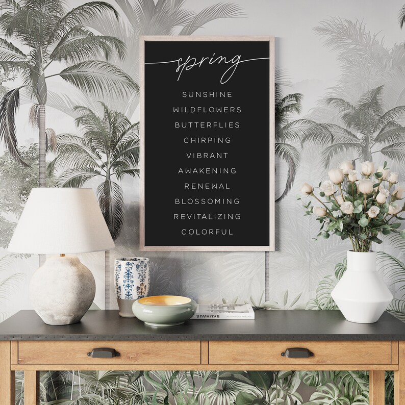 May include: A framed black print with the word "spring" in cursive script. Below, a list of spring-themed words is displayed in a clean, sans-serif font. The print hangs on a wall with a tropical palm tree wallpaper.