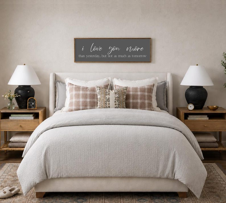 Puede incluir: Una escena de dormitorio con una cama con un edred&oacute;n gris claro y almohadas decorativas. Un letrero de madera sobre la cama dice "I love you more than yesterday, but not as much as tomorrow." Dos mesitas de noche con l&aacute;mparas completan la escena.