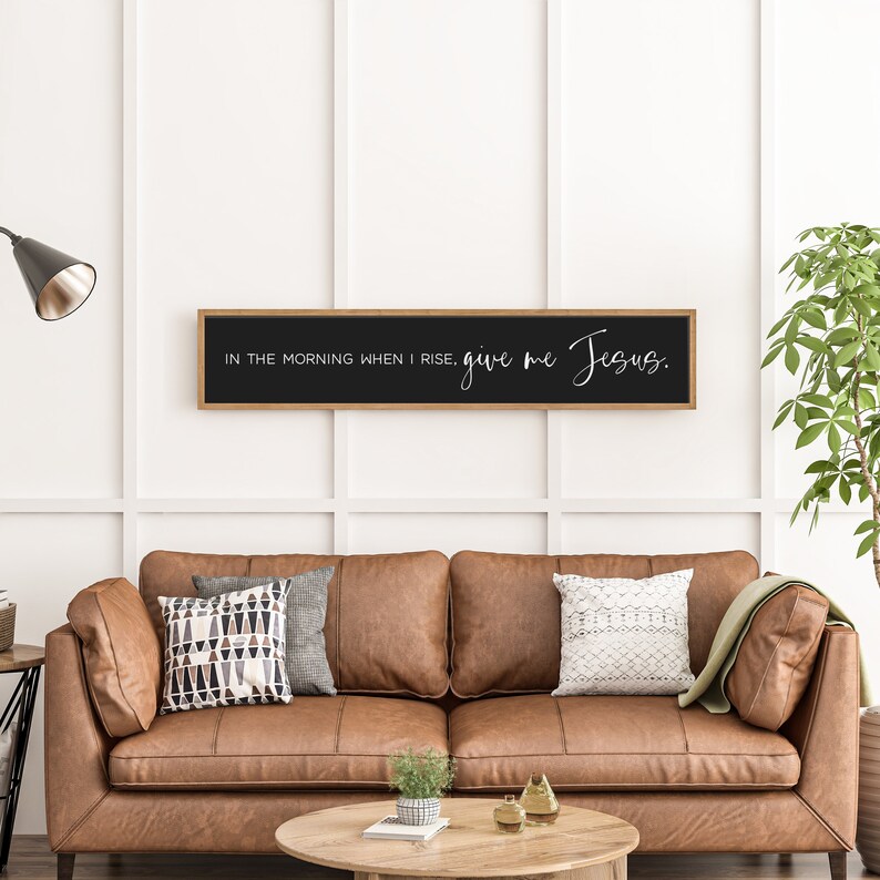 May include: A black framed sign with the text "In the morning when I rise, give me Jesus." hanging on a white wall above a brown leather couch.