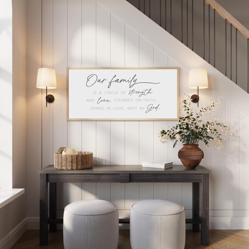 May include: A rectangular wall sign with a light wood frame featuring black text that reads: Our family IS A CIRCLE OF strength AND love. FOUNDED ON FAITH, JOINED IN LOVE, KEPT BY God. The sign is displayed on a white paneled wall above a dark console table.