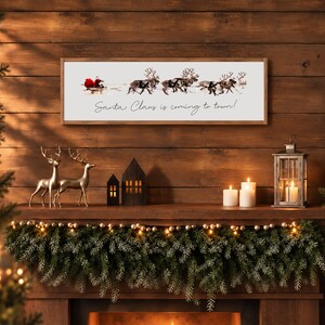 Puede incluir: Un letrero de pared rectangular con marco de madera y fondo blanco, que presenta un gr&aacute;fico de Santa Claus en un trineo tirado por renos y el texto cursivo negro Santa Claus is coming to town! centrado bajo la ilustraci&oacute;n.