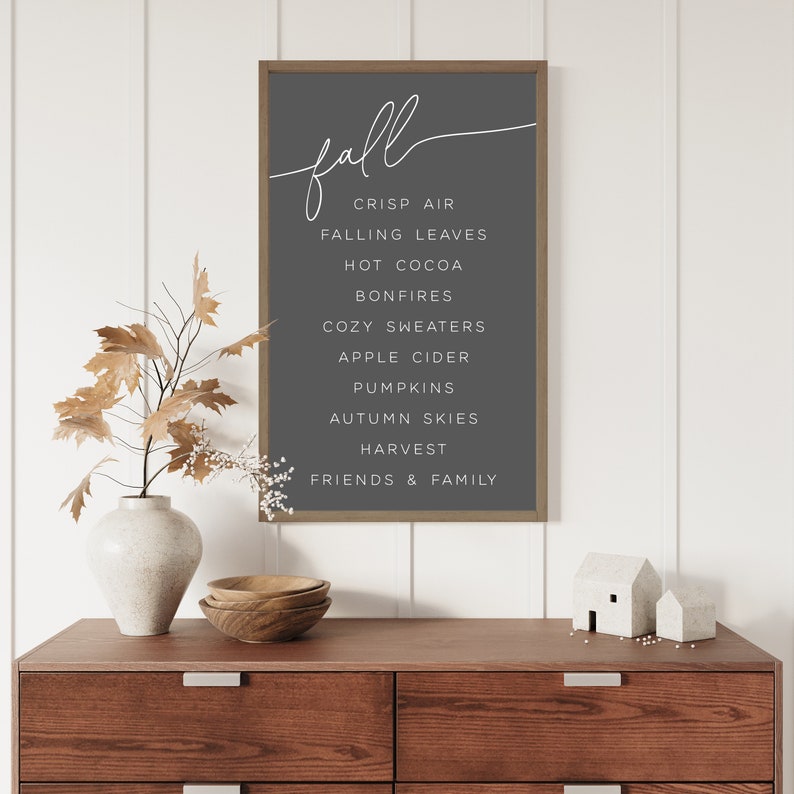 Op de afbeelding: Een ingelijst print met het woord "fall" in cursief schrift en een lijst met herfstgerelateerde woorden, waaronder "frisse lucht", "vallende bladeren", "warme chocolademelk", "vuurkorven", "gezellige truien", "appelcider", "pompoenen", "herfstlucht", "oogst" en "vrienden en familie". De print hangt aan een witte muur boven een houten dressoir.