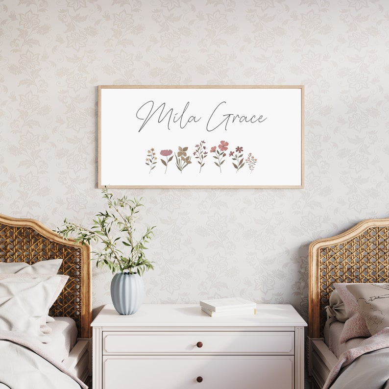May include: A rectangular wall sign in a light wood frame featuring the name Mila Grace in black cursive script above a row of small, muted-coloured botanical illustrations including various flowers and leaves on a white background.