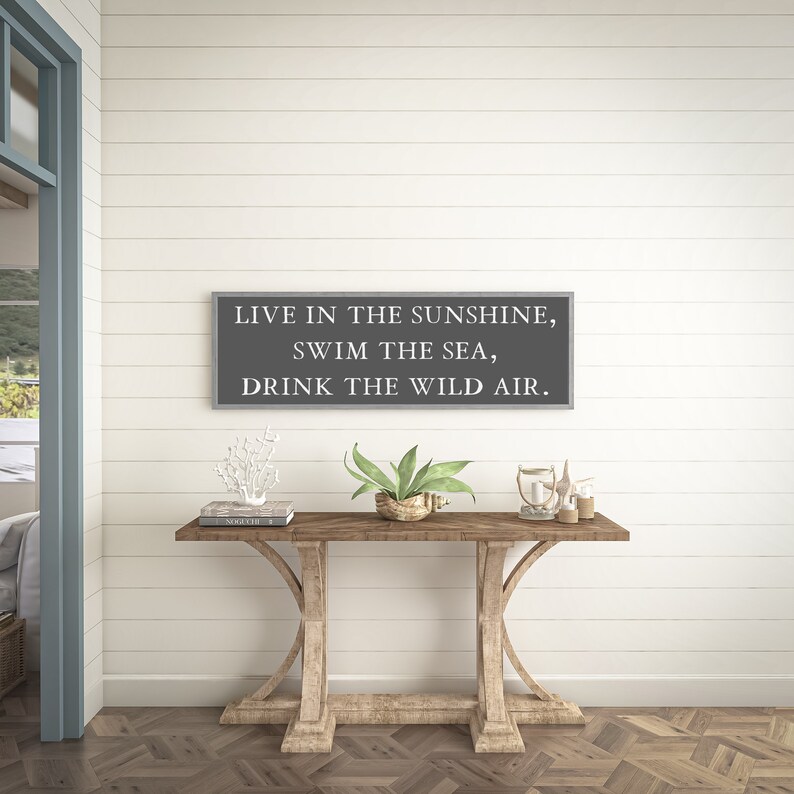 May include: A gray wooden sign with white text that reads "Live in the sunshine, swim the sea, drink the wild air." The sign is hanging on a white shiplap wall above a wooden console table with a plant, a book, and decorative items.