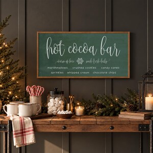 May include: A rectangular wooden sign with a dark green background and a light wood frame. The text reads "hot cocoa bar" in large script, with "warm up here" and "made fresh daily" flanking a snowflake, and a list of toppings below.