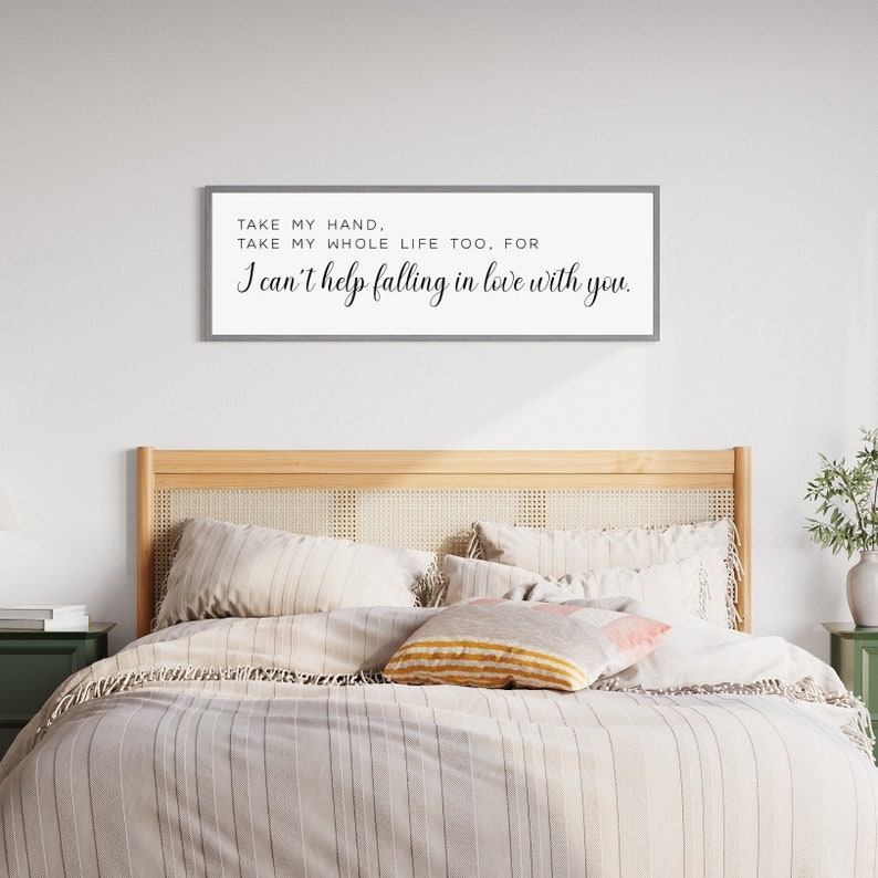 Op de afbeelding: Een rechthoekig, ingelijst bord met de tekst "TAKE MY HAND, TAKE MY WHOLE LIFE TOO, FOR I can't help falling in love with you." wordt weergegeven boven een bed met neutraal gekleurd beddengoed en kussens. Het bed heeft een licht houten hoofdbord.
