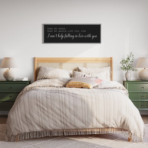 Op de afbeelding: Een slaapkamer met een bed met een lichtgekleurde gestreepte dekbedovertrek en decoratieve kussens. Boven het bed hangt een zwart kader met witte tekst: "TAKE MY HAND. TAKE MY WHOLE LIFE TOO, FOR I can't help falling in love with you."