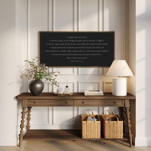 May include: A wooden console table with a dark brown frame, two drawers, and a lower shelf. Two woven baskets sit below. A framed black sign with white text hangs above the table. A vase of flowers and a lamp are also on the table.