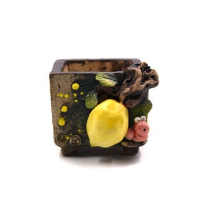 May include: A square, ceramic planter with a textured, mottled finish. The planter is decorated with a large yellow lemon, green leaves, and a pink worm-like creature. The planter is suitable for small plants.