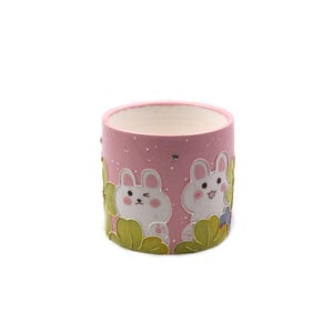 Puede incluir: Una maceta de cerámica rosa con interior blanco. El exterior presenta dos conejos de dibujos animados blancos con mejillas rosas y follaje verde. La maceta es cilíndrica y tiene una superficie ligeramente texturizada.