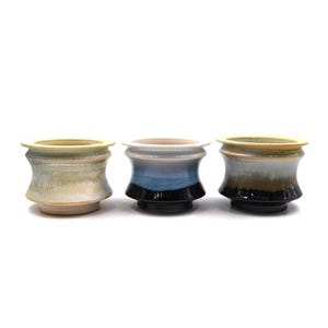 May include: Three ceramic cups with a flared rim and a tapered base. The left cup features a pale yellow and green glaze. The center cup has a blue and black gradient glaze. The right cup displays a grey, brown, and black blended glaze finish.