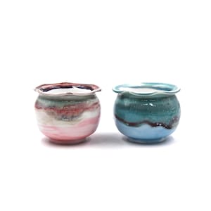 May include: Two small, round ceramic pots with ruffled rims. One pot features pink, white, and red hues, while the other is teal and blue. Both have a glossy finish and are suitable for small plants or decorative use.