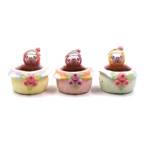 Peut inclure: Trois pots de fleurs en céramique, chacun représentant un paresseux portant un chapeau. Les pots sont de couleurs pastel : jaune, rose et vert clair. Chaque pot est décoré d'un motif floral et de pois.