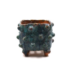 May include: A square ceramic planter with a textured, bumpy surface in shades of deep blue, teal, and green. The vessel features a glossy, mottled glaze finish and stands on four short, sturdy feet, with a warm brown clay tone visible at the base.